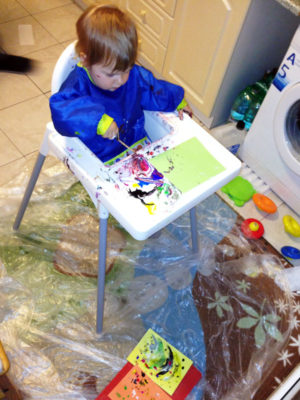 Ako začať s deťmi s maľovaním - dieťa sedí na vysokej stoličke, pod sebou má igelit, sa sebe ochrannú zásterku a maľuje farbami na papier